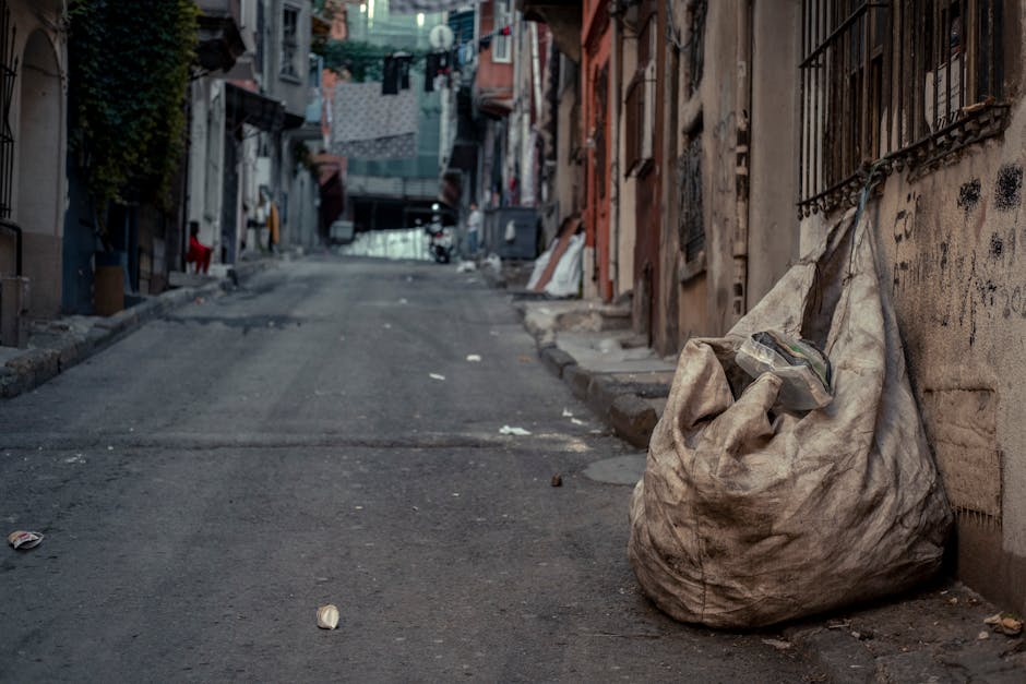 A large, worn and dirty white garbage bag, made of heavy-duty plastic material, is positioned on a narrow, uneven urban street pavement next to a weathered brick wall with iron security bars. The bag