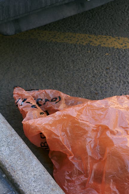 A large, worn and dirty white garbage bag, made of heavy-duty plastic material, is positioned on a narrow, uneven urban street pavement next to a weathered brick wall with iron security bars. The bag